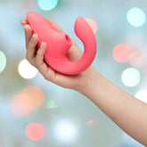 A hand holding a vibrant rose Womanizer Blend Air-Pulse Clit Stimulator against a softly blurred background with pastel-colored circular bokeh lights. The toy features an ergonomically curved design and a rounded tip designed for blended orgasms.